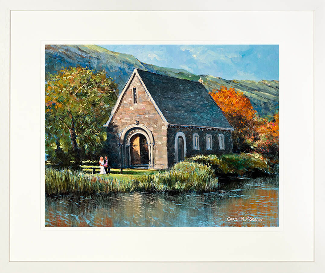 A framed print of a painting of a wedding couple sitting on a seat in front of the chapel at Gougane Barra, County Cork