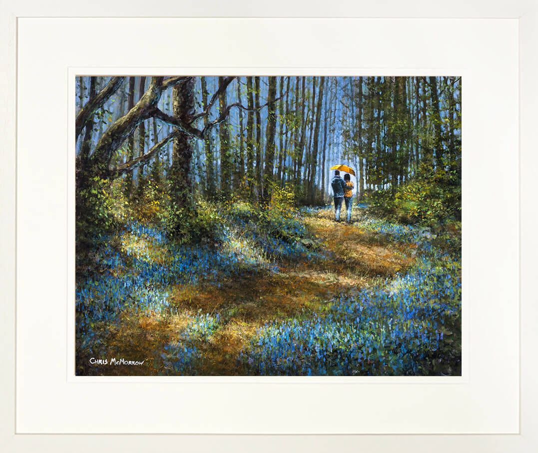 A framed print of a painting with a couple linking under an umbrella walking through a path of bluebells in a wood